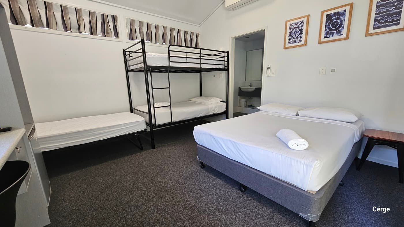 The ensuite room contains a single bed and a double-decker bed on the left, with a double bed adjacent. All beds are covered with white sheets. A small table is positioned on the right of the bed. Picture frames decorate the upper right wall. The floor has a dark grey carpet. The ceiling is white, and the left wall has short curtains. The toilet is positioned in the right corner.