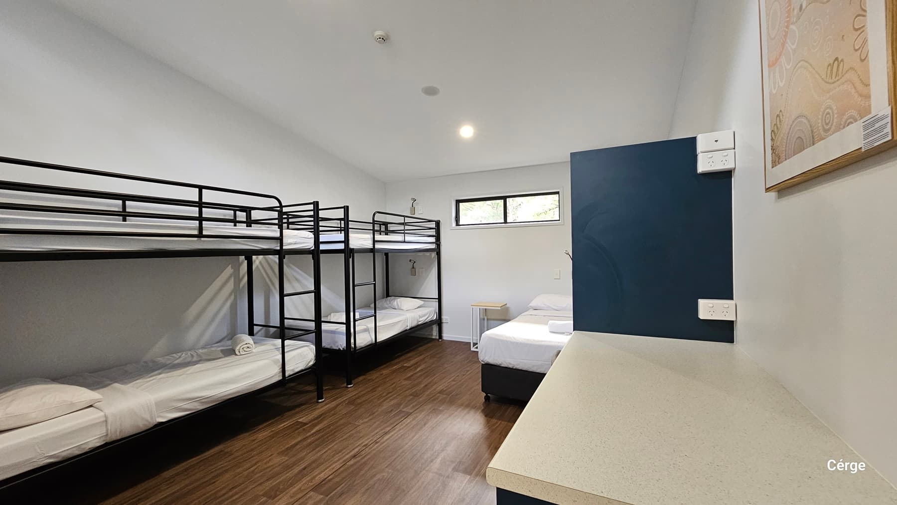 On the left, there are two sets of black metal bunk beds with white bedding. On the right, a blue door is partially open, revealing a single bed with white bedding. Above the bed, a small rectangular window lets in light. In the foreground on the right, a long white countertop extends along the wall. The floor is made of dark timber, and the ceiling is plain white with recessed lighting.