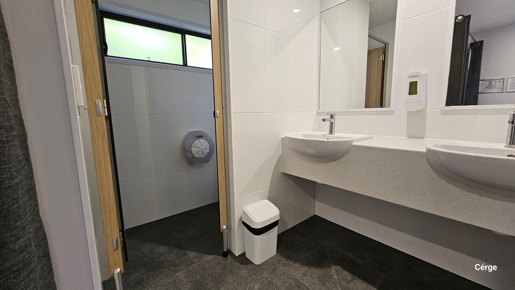 On the left, there is a partially open door revealing a toilet cubicle with a toilet paper dispenser mounted on the wall. The floor is dark grey tile. On the right, there are two white sinks with a soap dispenser mounted on the wall above them. A large mirror is positioned above the sinks. The background features white tiled walls. The ceiling is white and smooth. In the foreground, there is a small white bin placed under the sinks.