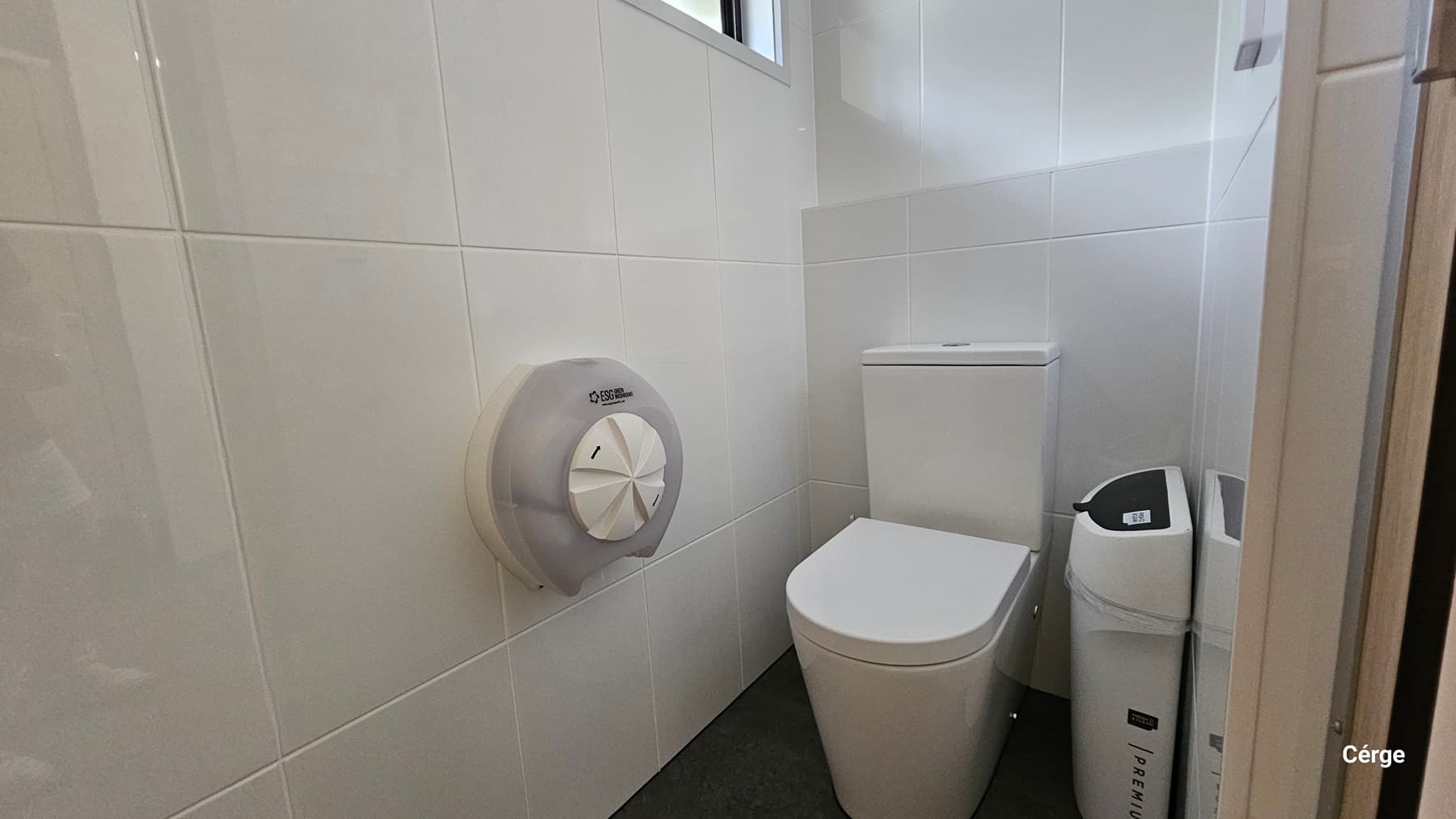 A small bathroom with white tiled walls. On the left, there is a large, round toilet paper dispenser mounted on the wall. In the middle, there is a white toilet with a rectangular cistern. To the right of the toilet, there is a white bin with a black lid. The floor is covered with dark grey tiles.