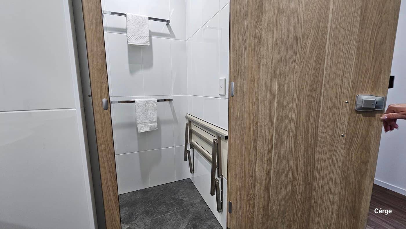 A small bathroom with white tiled walls. On the left, there is a white towel hanging on a metal rail. On the right, a wooden door is partially open. In the middle, there is a foldable shower bench attached to the wall. The floor is covered with dark grey tiles.