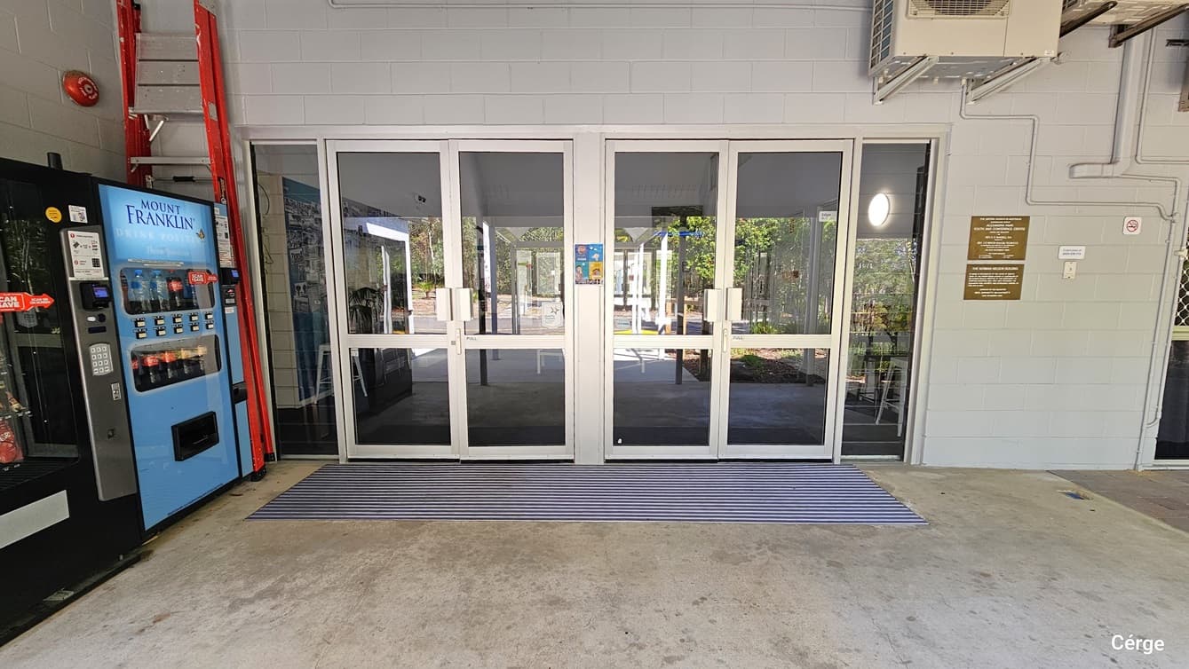 A pair of double glass doors set in a light grey wall within a building, flanked by red fire extinguisher equipment to the left and a vending machine for cold drinks. An orange ladder is placed against the wall.  A striped mat lies in front of the doors. Signs and notices are visible on the wall next to the door, and the interior behind the glass doors appears to be a foyer or entrance hall with seating.