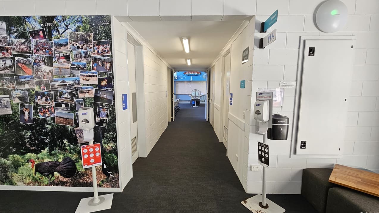 The corridor is carpeted in a dark color and lined with white walls. On the left wall is a large collage of photographs pinned close together, featuring images of people and activities. Hand sanitizing dispensers are placed on both sides.  Toilet signage is placed on both sides of the wall. An open door leads into a bright, blue-coloured room at the far end of the corridor.