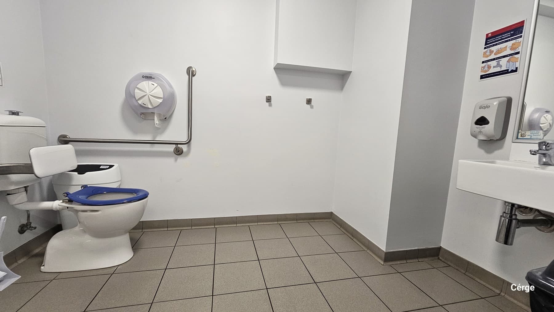 An accessible toilet featuring a wash basin on the right. Paper towel and soap dispensers are mounted to the wall, along with a sign. A black rubbish bin is placed under the wash basin. In the left corner is a toilet bowl with a blue seat cover, a rubbish bin, and grab bars. The wall is all white, and the floor has grey tiles.