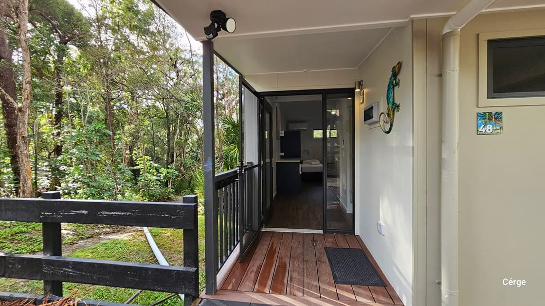 On the left, there is a black wooden railing with a view of a green, wooded area with various trees and plants. On the right, a beige wall with a colourful gecko decoration and a room number sign "48". In the middle, a glass sliding door leads to an interior space with a visible bed. The foreground features a wooden deck floor with a black doormat. The ceiling is white and has a light fixture.