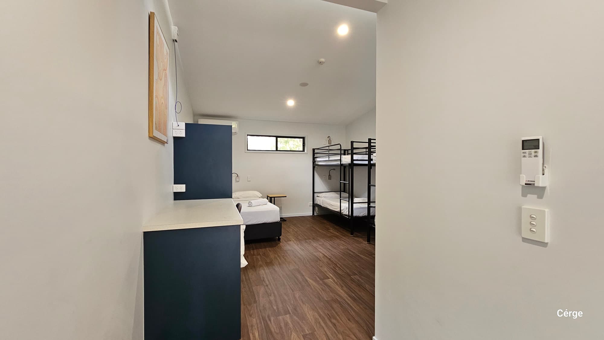 A room with a timber floor and white ceiling with recessed lighting. On the left, there is a blue and white cabinet with a framed photo above it. In the middle, a single bed with white bedding is positioned against the wall. On the right, there are two black metal bunk beds. At the end, there's a rectangular window with a view of greenery. The foreground includes a white wall with a thermostat and light switch.
