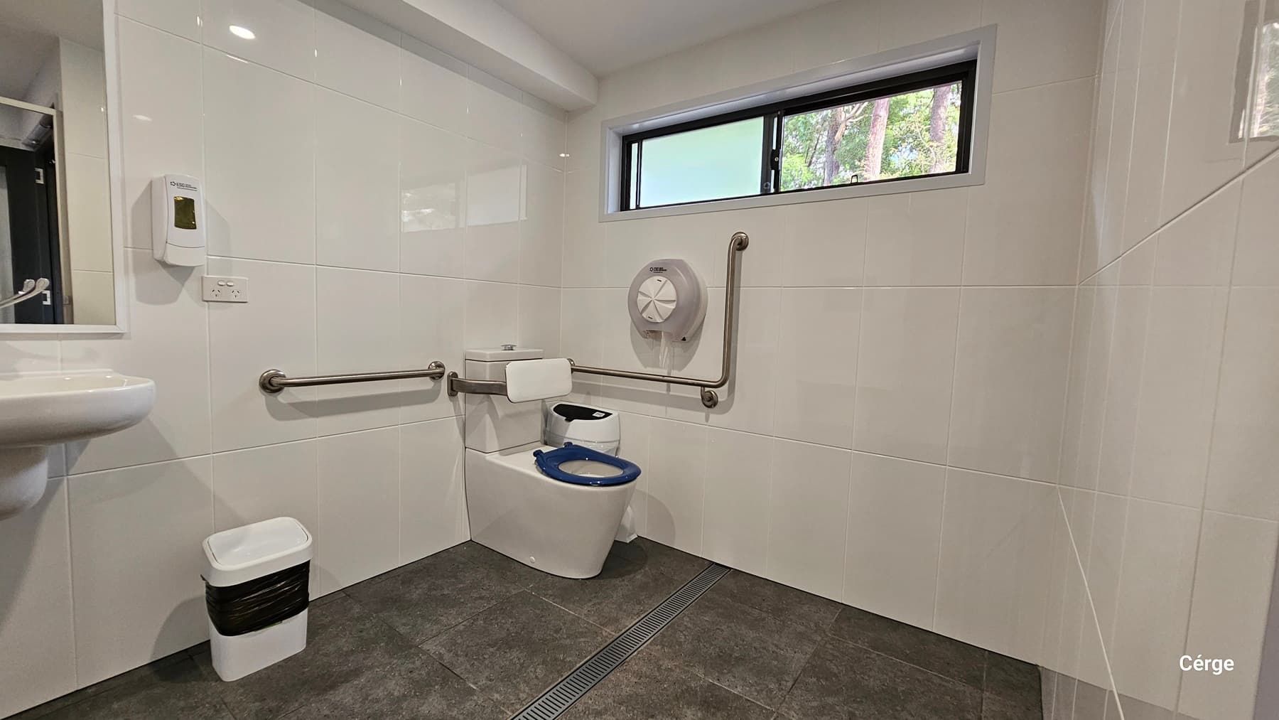 On the left, there is a white sink with a soap dispenser mounted on the wall above it. Below the sink, a small white bin with a black liner is placed. In the middle, a white toilet with a blue seat is installed, accompanied by stainless steel grab bars on both sides. On the right, a large toilet paper dispenser is mounted on the wall. The floor is covered with dark grey tiles, and the walls are lined with white tiles. A rectangular window is positioned high on the wall.