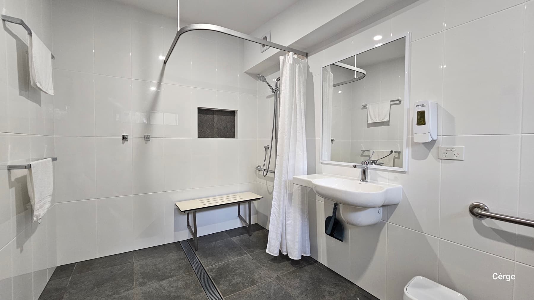 A bathroom with white tiled walls and a dark grey tiled floor. On the left, there are two towel racks with white towels. In the middle, a shower area with a white curtain, a wall-mounted showerhead, and a foldable bench. On the right, a white sink with a mirror above it, a soap dispenser, and a grab bar. The ceiling is white with recessed lighting.