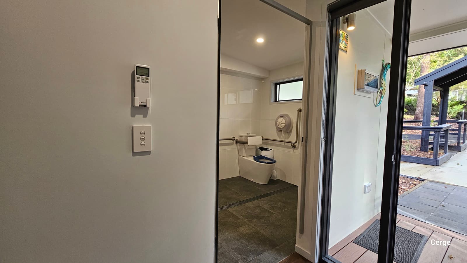 On the left is a white wall with a thermostat and a light switch. In the middle, is a bathroom with a toilet, grab bars, and a small window above. The floor is dark grey tiles. On the right, a glass door leads outside to a patio area with a black railing and a pathway. The ceiling is white with recessed lighting.