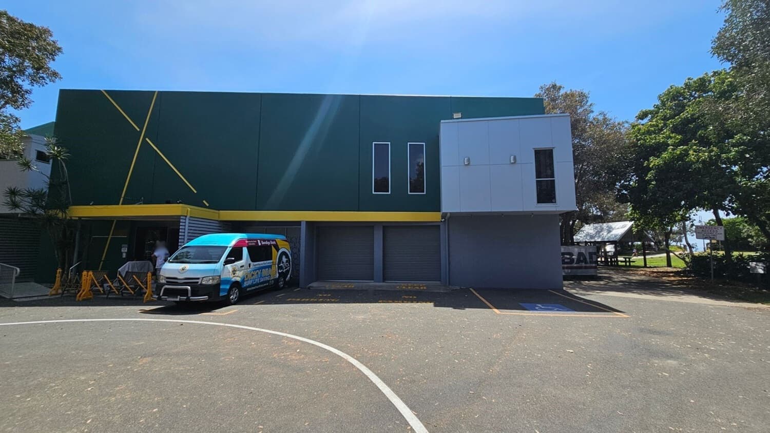 A two-storey building with a dark green wall on the left featuring yellow diagonal lines and a yellow trim above the ground level. On the right, there is a grey panelled section with a single window. At ground level, three grey roller doors are visible. In front of the left section, a white van with colourful branding is parked near bright yellow barriers. The foreground features a tarmac surface with parking markings. Trees and a small shelter are in the background on the right.