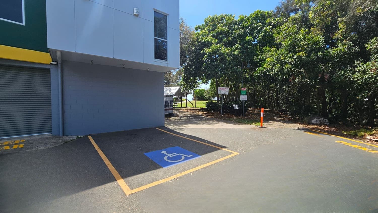 On the left, a building with a grey roller door and a yellow trim sits adjacent to a grey wall with a window above. On the right, dense green trees line the edge of the area, with signs and a bright orange bollard near a dirt pathway. In the middle, there is a marked blue disabled parking space surrounded by yellow boundary lines. The floor is asphalt, partially shaded, with a smooth, slightly worn texture.
