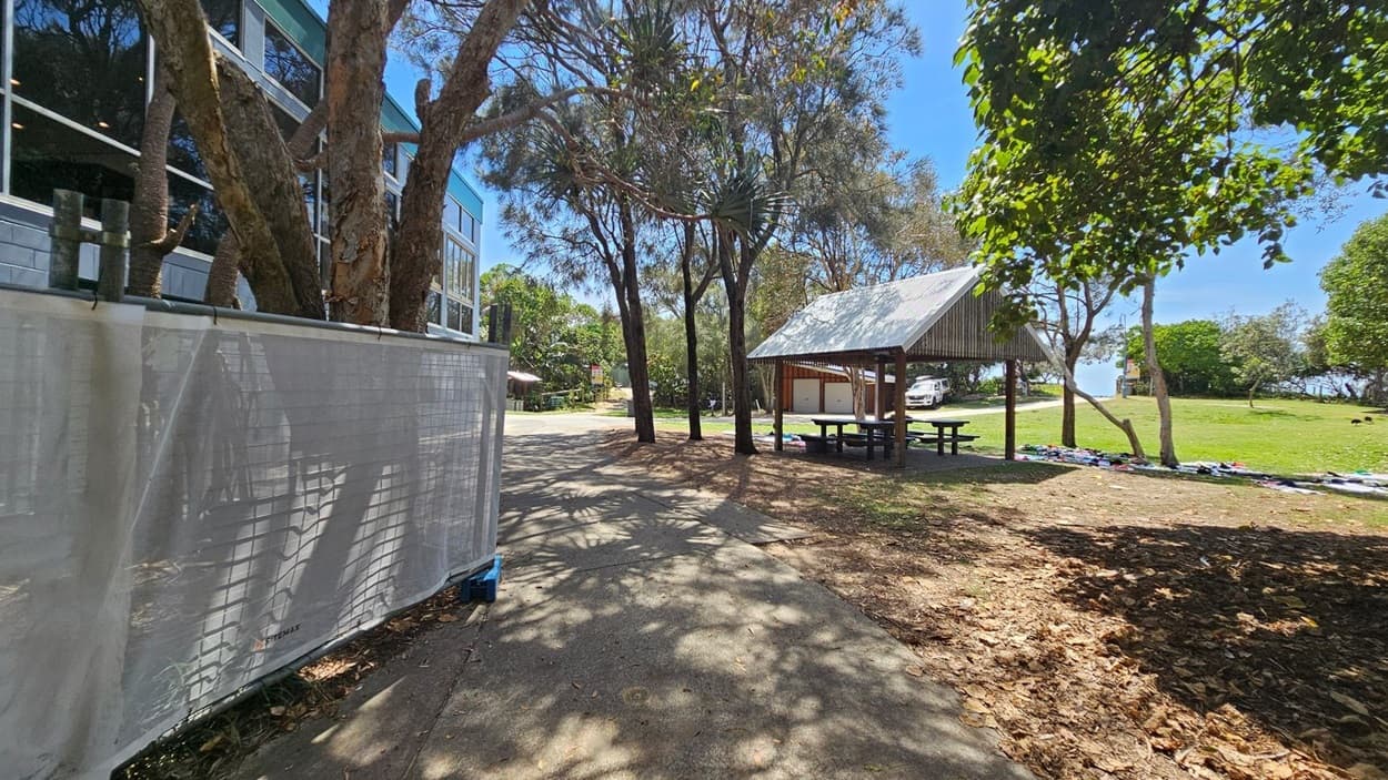 On the left, a multi-storey building with large windows is partially obscured by several trees and a white mesh barrier. On the right, a timber picnic shelter with a metal roof stands next to scattered trees and an open grassy area. In the foreground, a concrete pathway extends through dappled shade from surrounding trees, while dry leaves are scattered on the ground. A white vehicle is visible in the background near more trees and shrubs.