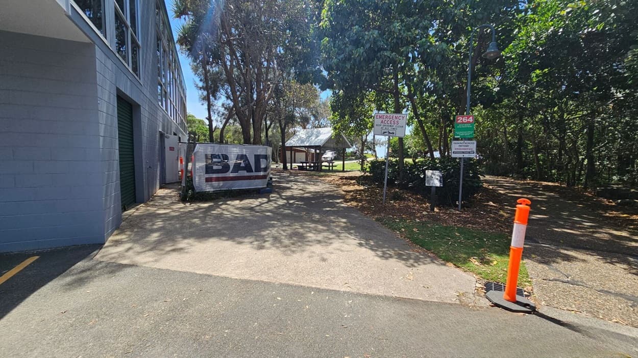 On the left, a grey building with large windows and two green roller doors extends along the edge of the image. In the middle, a concrete pathway leads to a picnic shelter with a metal roof and wooden picnic tables, surrounded by trees and grass. On the right, signs, an orange bollard, and a streetlamp are near a gravel pathway with dry leaves scattered. The floor consists of asphalt transitioning to concrete and gravel.