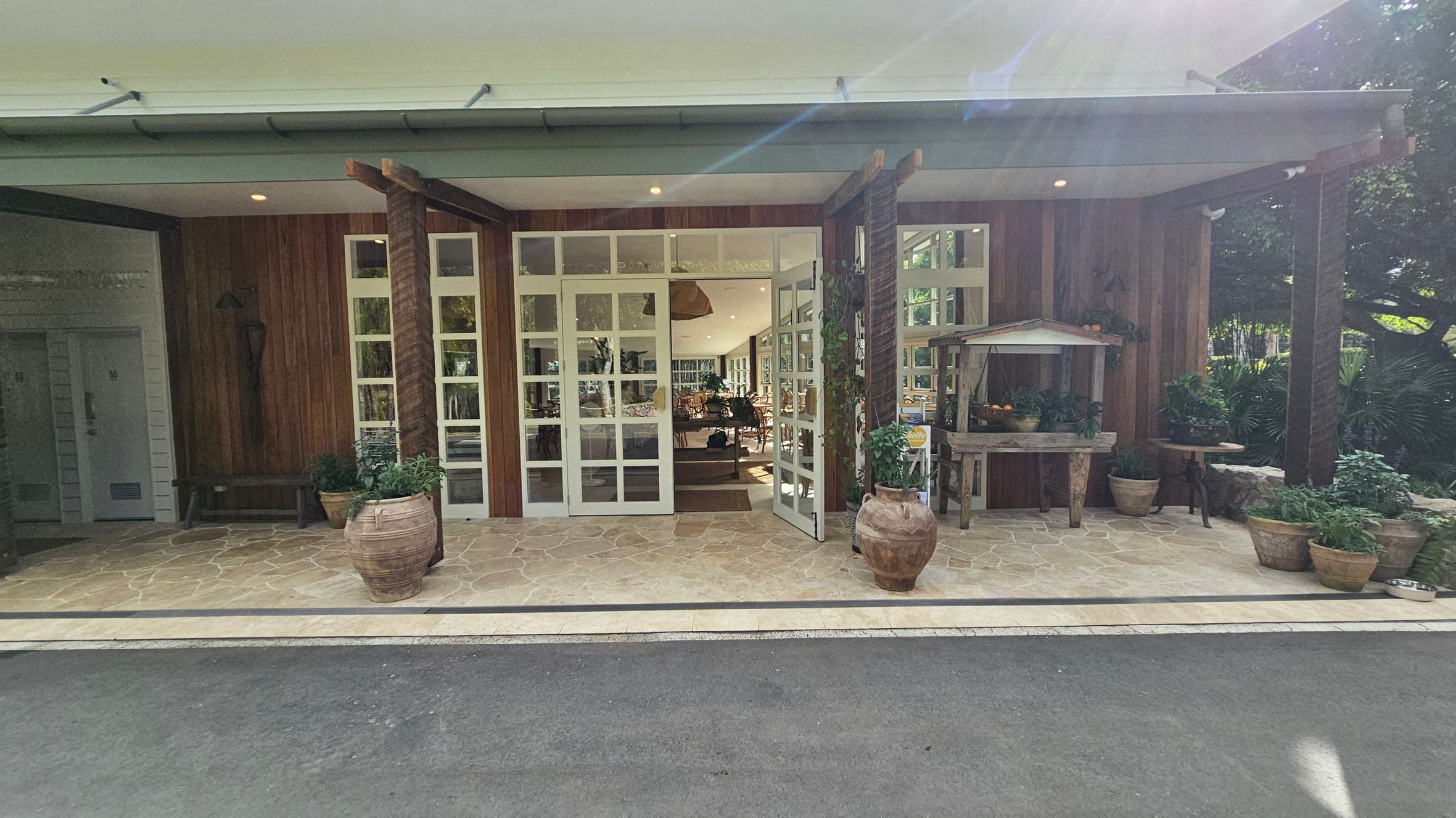 A building entrance with large glass doors in the centre, flanked by wooden beams. The floor is paved with stone tiles. On the left, there's a wooden wall with a bench and potted plants. On the right, a small wooden table holds various items, surrounded by more potted plants. The ceiling is white with recessed lighting. Inside, tables and chairs are visible, suggesting a dining area.
