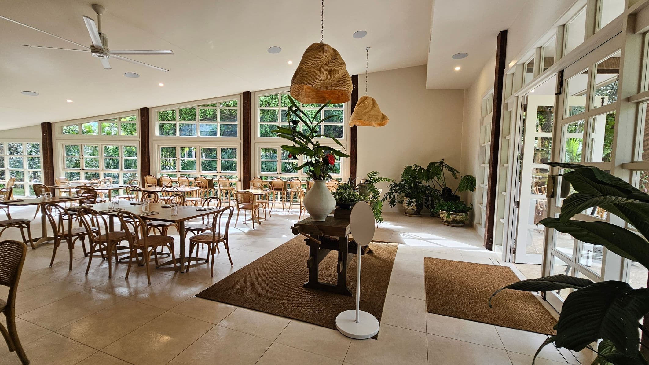 A spacious indoor dining area with large windows on the left, allowing natural light to flood in. Wooden tables and chairs are arranged neatly, with some tables set for dining. In the centre, a wooden table holds a large vase with green plants. Woven pendant lights hang from the ceiling. On the right, glass doors open to an outdoor area, and several potted plants are placed around. The floor is tiled, and there are brown rugs near the entrance.