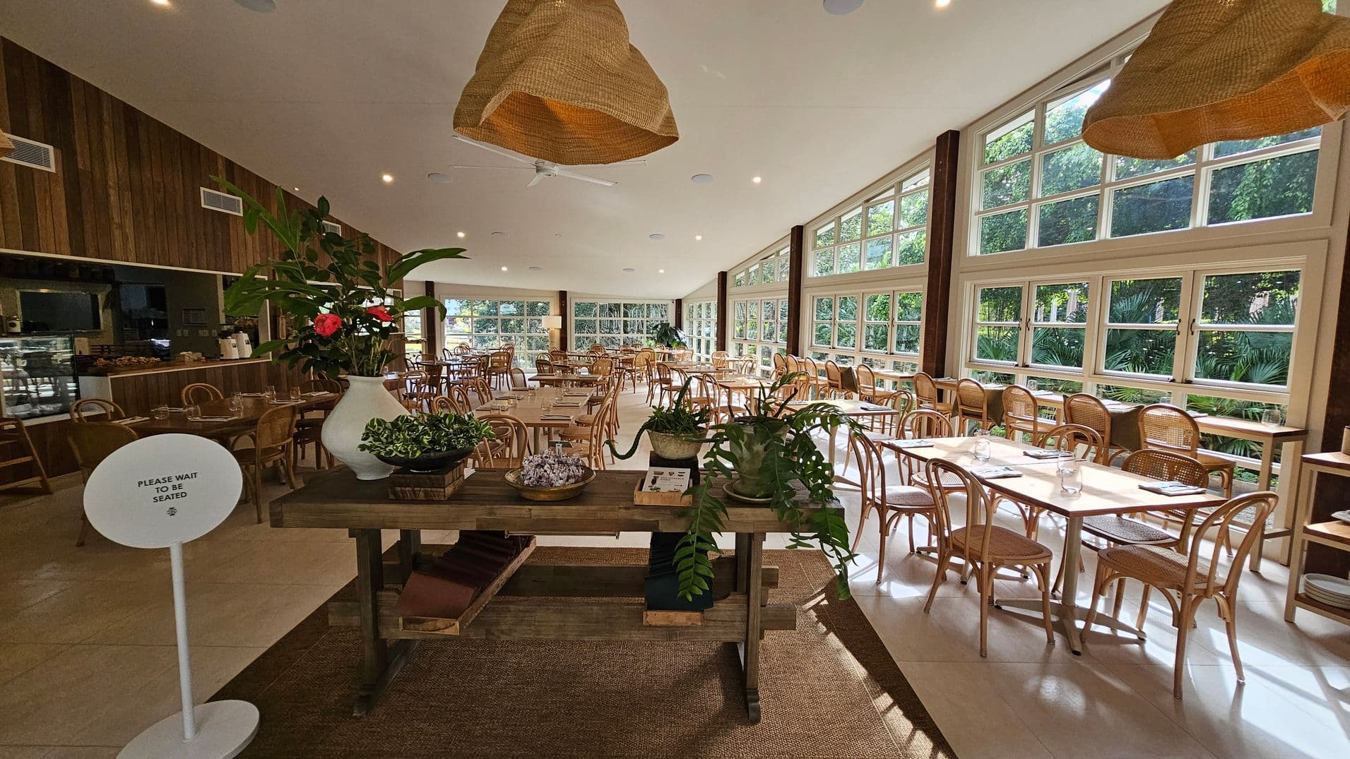 A spacious dining area with large windows on the right, allowing natural light to illuminate the room. Wooden tables and chairs are neatly arranged throughout. In the foreground, a wooden table displays potted plants and a sign that reads "Please wait to be seated." The ceiling features woven pendant lights, and the floor is a mix of tiles and a woven rug. The left side has a wooden wall with a counter and more seating. Lush greenery is visible outside the windows.