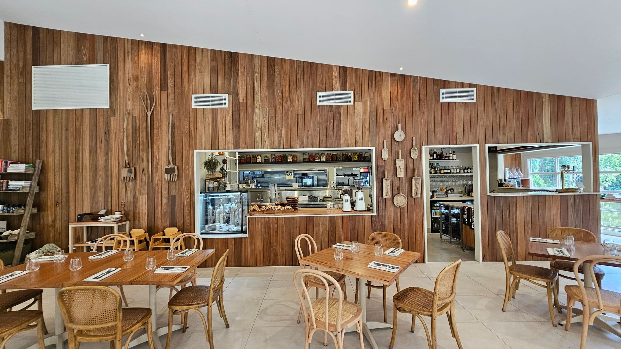 A cosy café interior with wooden panelled walls. In the foreground, wooden tables are set with glassware and napkins, surrounded by wicker chairs. On the left, shelves hold books and items, with a ladder leaning against them. The middle features a window into the kitchen, displaying jars and kitchen equipment. On the right, a small open doorway reveals more kitchen supplies. The floor is tiled, and the ceiling is plain white.