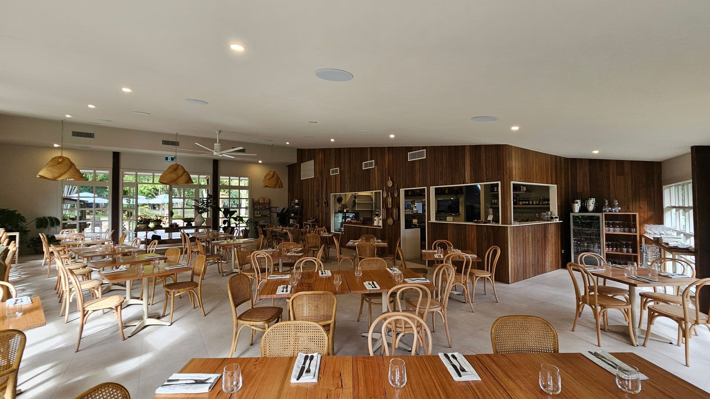 Spacious restaurant interior with wooden tables and wicker chairs arranged neatly. The left side features large windows allowing natural light, with potted plants adding greenery. The right side has a wooden counter with shelves displaying bottles and glassware. The ceiling is light-coloured with recessed lighting and a ceiling fan. The floor is light-coloured, complementing the warm wooden tones throughout the space.