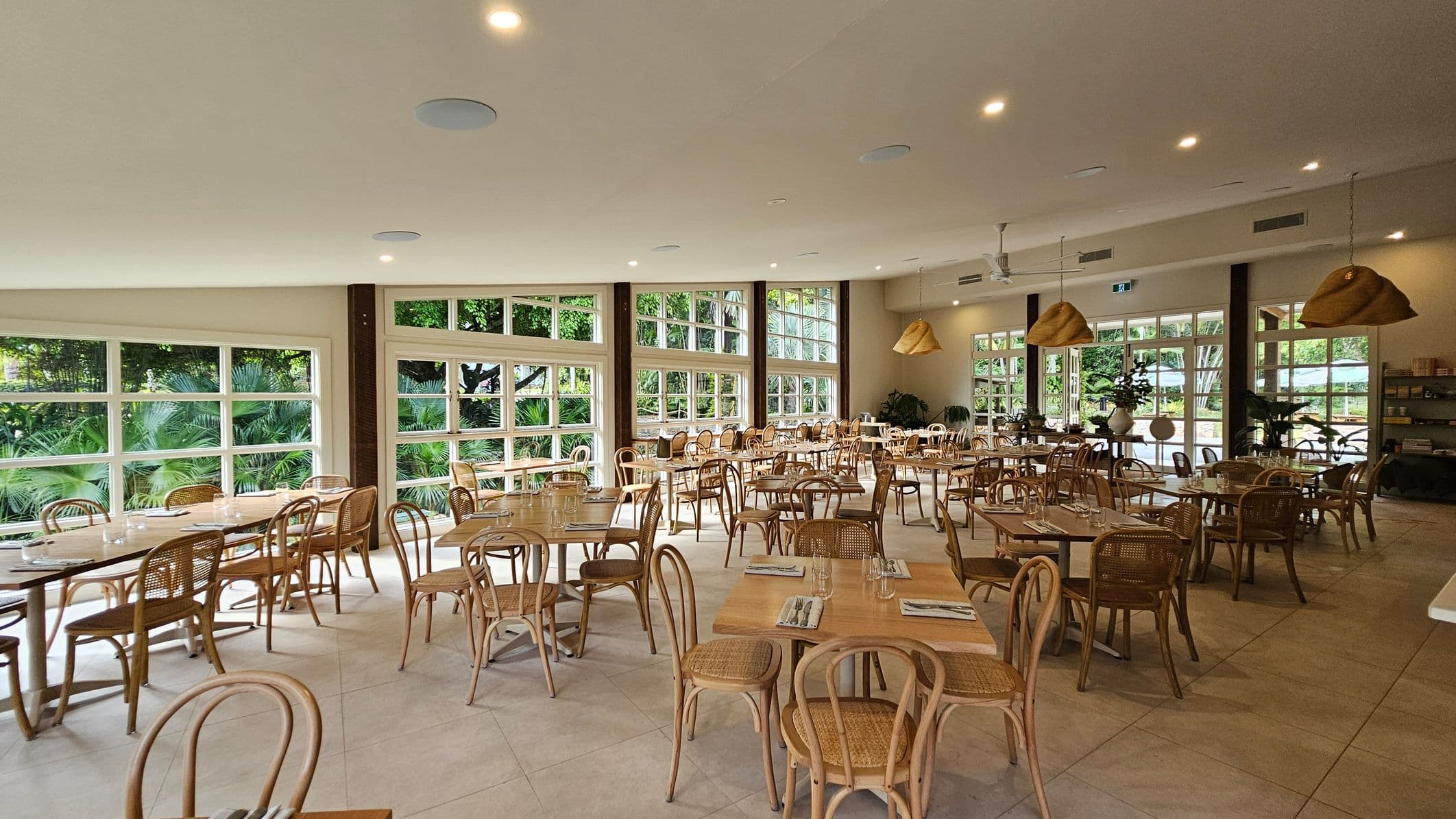 Spacious dining area with wooden tables and chairs arranged neatly. Large windows on the left and back walls allow natural light to flood in, offering views of lush greenery outside. The ceiling is fitted with recessed lights and features hanging woven lampshades. The floor is tiled in a light colour. In the background, there are potted plants and shelves with items. The overall atmosphere is airy and inviting.