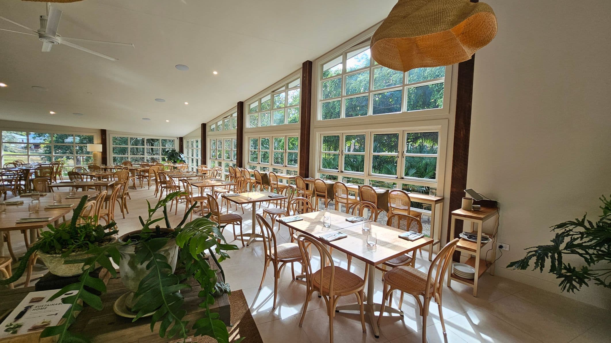 Spacious dining area with large windows on the right, allowing natural light to flood in. Wooden tables and chairs are neatly arranged throughout the room. A ceiling fan is visible above, and a woven lampshade hangs from the ceiling. In the foreground, there are potted plants on a table. The floor is light-coloured, complementing the bright and airy atmosphere.