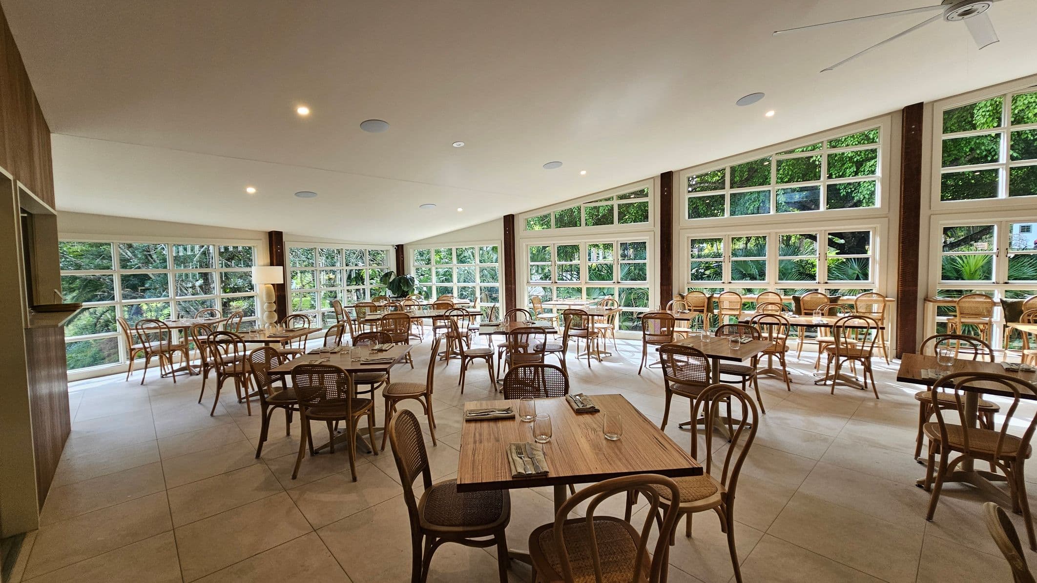 A spacious dining area with wooden tables and chairs arranged neatly across the room. Large windows on the right side let in natural light and offer a view of lush greenery outside. The ceiling is light-coloured with recessed lighting and a ceiling fan. The floor is tiled in a light shade. A tall lamp and a potted plant are near the windows, adding to the decor.