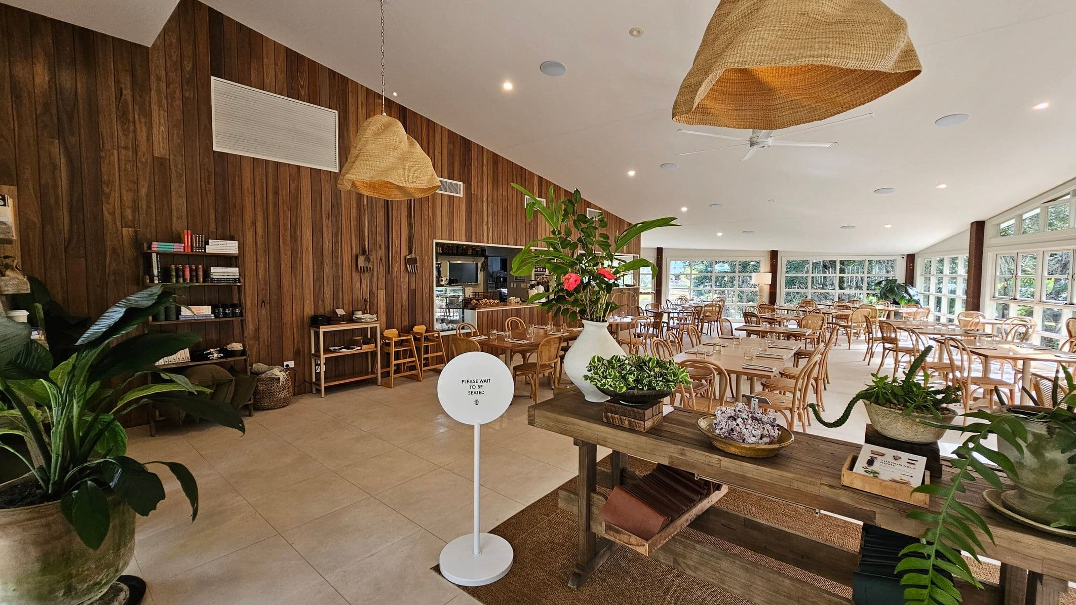 A spacious restaurant interior with a wooden wall on the left featuring shelves with books and decor. A small counter with stools is nearby. In the centre, a wooden table displays plants and a sign reading "Please wait to be seated." The right side has numerous wooden tables and chairs, with large windows allowing natural light. The ceiling is white with hanging woven lampshades, and the floor is tiled.
