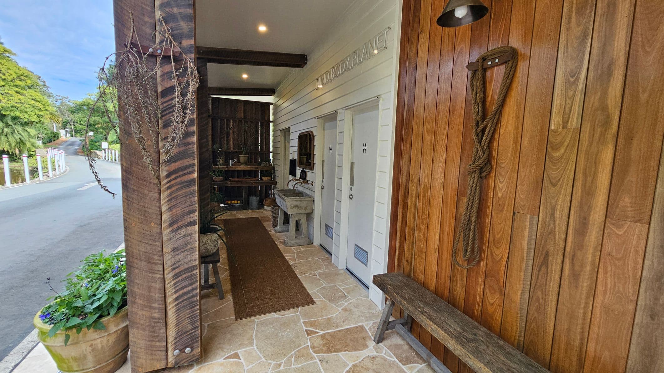 A narrow outdoor corridor with a stone-tiled floor. On the right, a wooden wall features a coiled rope and a bench. Two white doors with gender-neutral symbols are in the centre, next to a rustic sink. Above, a sign reads "MIND BODY PLANET." On the left, a large wooden post with hanging dried branches and a planter with green foliage. The background shows a road curving left, lined with white posts and surrounded by lush greenery. The ceiling has recessed lighting.