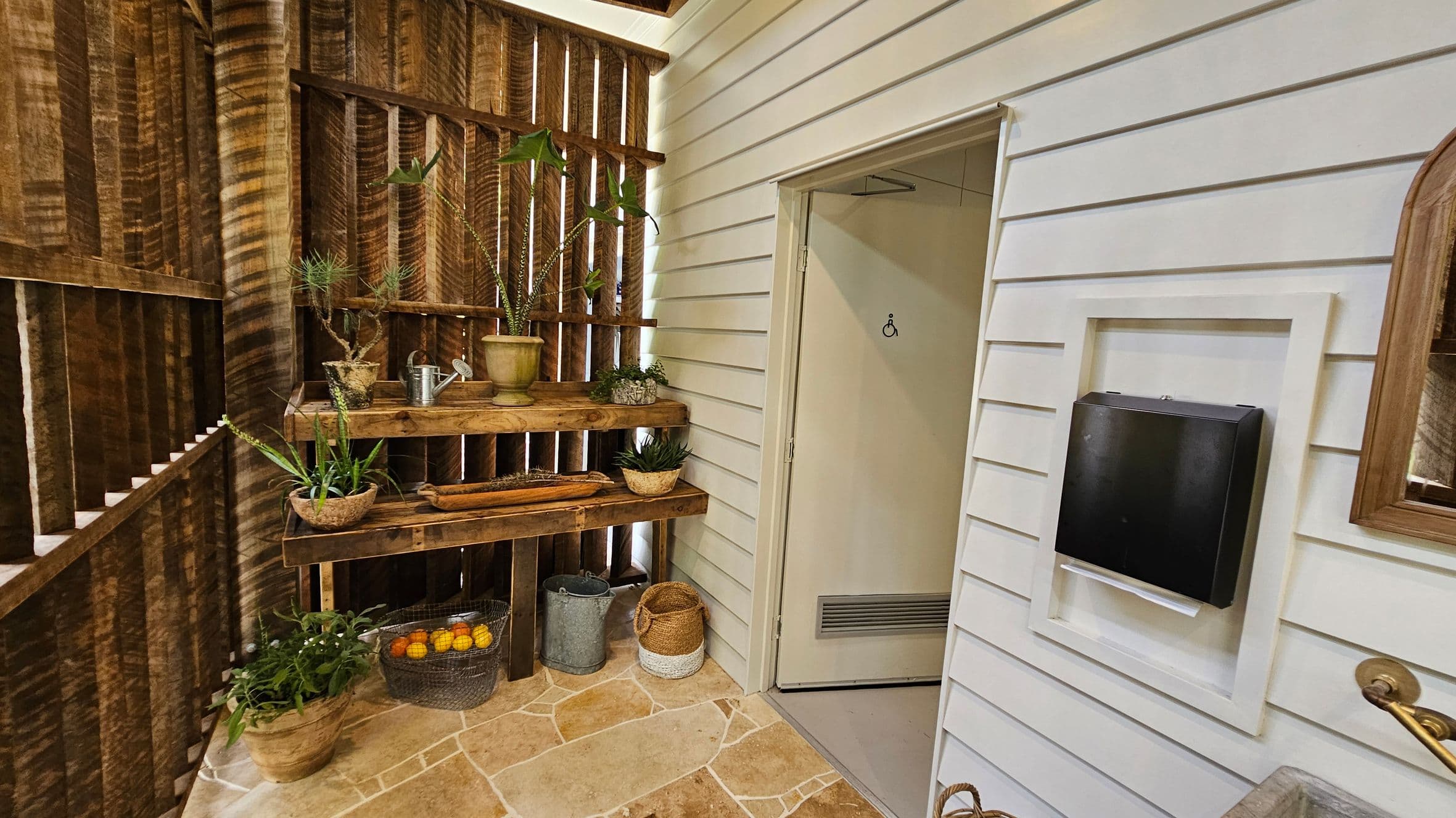 A rustic interior with a wooden wall on the left featuring vertical slats. A wooden shelf holds various potted plants and a watering can. Below, baskets contain oranges and other items. The floor is tiled with a stone pattern. On the right, a white panelled wall has a door with a disability sign. A black box is mounted on the wall, framed in white. A mirror with a wooden frame is partially visible.