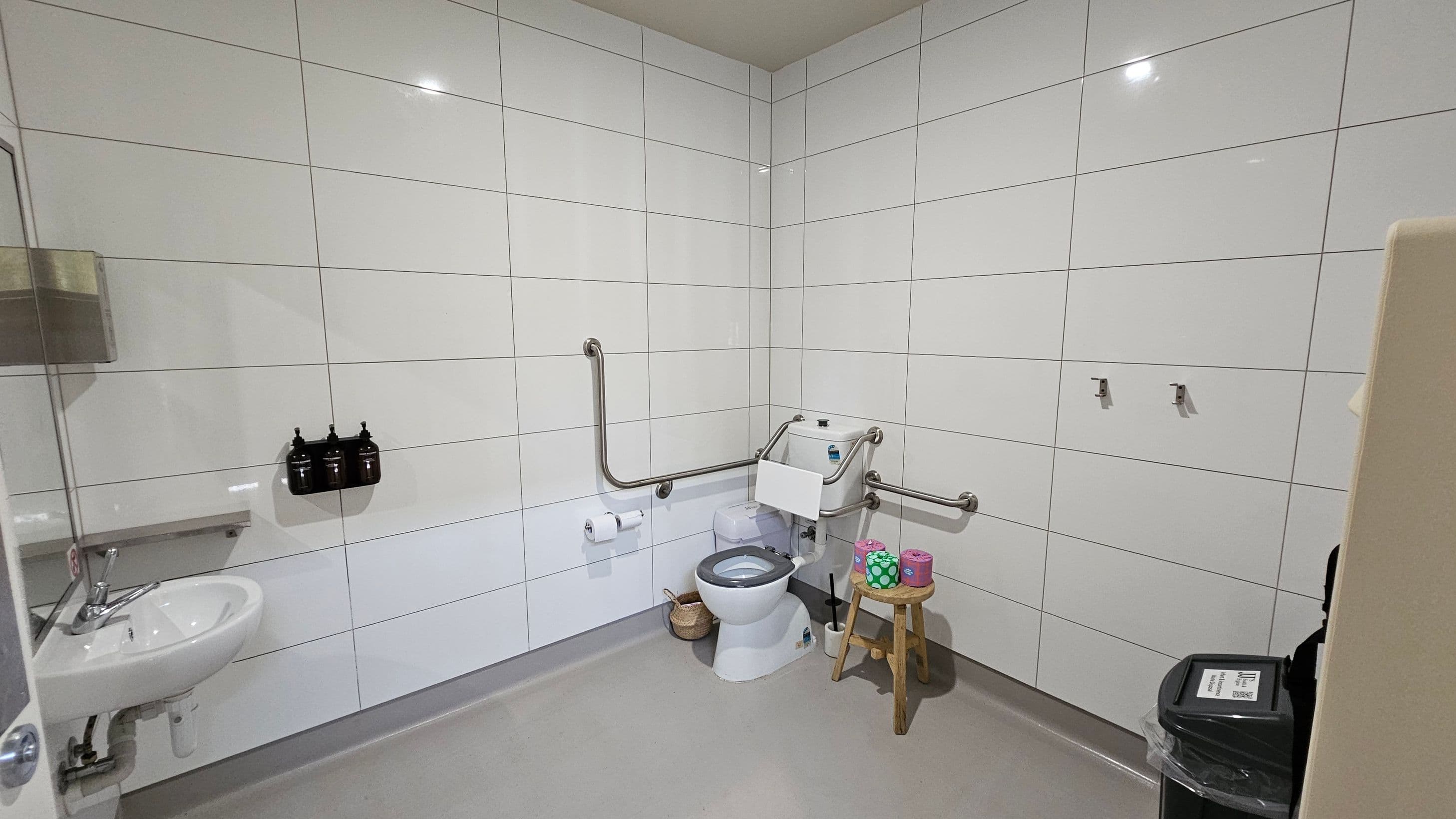 A spacious bathroom with white tiled walls and a grey floor. On the left, a sink with a metal tap is mounted on the wall, accompanied by a soap dispenser. In the centre, a toilet with support rails is positioned, with a small wooden stool nearby holding colourful toilet rolls. A basket is placed on the floor beside the toilet. On the right, a black bin with a lid is visible. The ceiling is light green, and there are two hooks on the wall near the toilet.