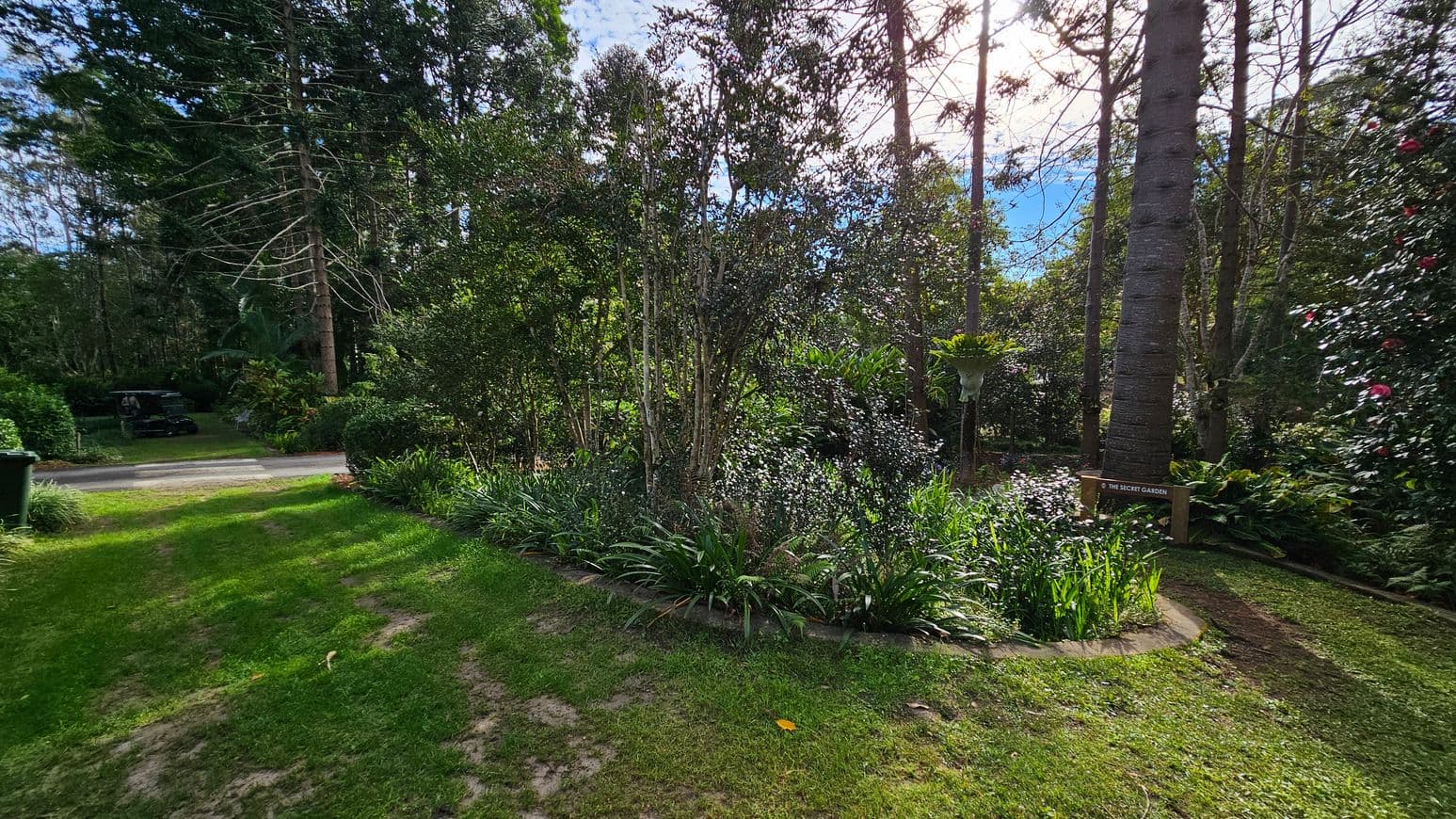 A lush garden scene with a variety of trees and shrubs. In the centre, a circular garden bed is filled with dense foliage and tall plants. To the right, a sign reads "The Secret Garden" near a large tree trunk. The background is filled with more trees, creating a dense canopy. On the left, a path leads to a small vehicle partially visible among the greenery. The grass in the foreground is well-maintained, with patches of sunlight filtering through the trees.