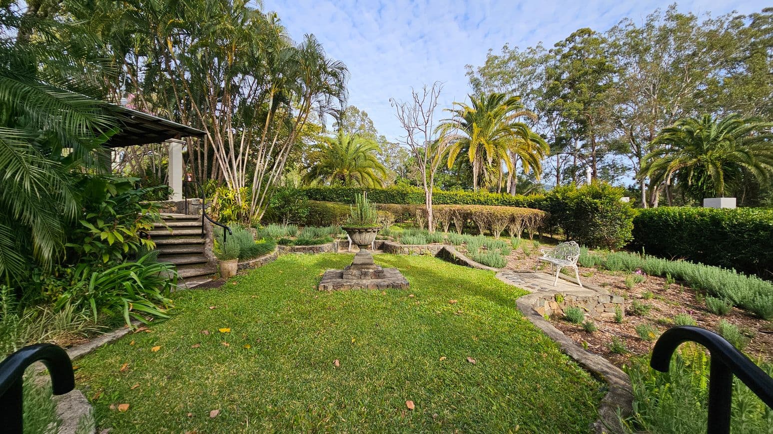 A lush garden with a central stone planter filled with greenery. On the left, a set of stone steps leads up to a covered area surrounded by dense foliage and tall palm trees. On the right, a white metal bench sits on a stone path amidst low shrubs and plants. The background features a hedge and more palm trees, with taller trees beyond. The foreground is a well-maintained grassy area with a few scattered leaves.