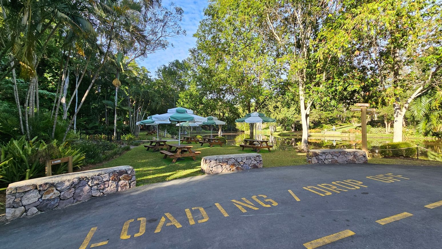 A park area with several wooden picnic tables and green-and-white umbrellas on a grassy lawn. In the foreground, a paved area marked with "LOADING / DROP OFF" in yellow letters. Stone walls border the paved area. Lush greenery, including tall trees and palm trees, surrounds the space. A small pond is visible in the background, reflecting the trees. A wooden signpost stands near the pond, and a variety of plants and shrubs are scattered throughout the scene.