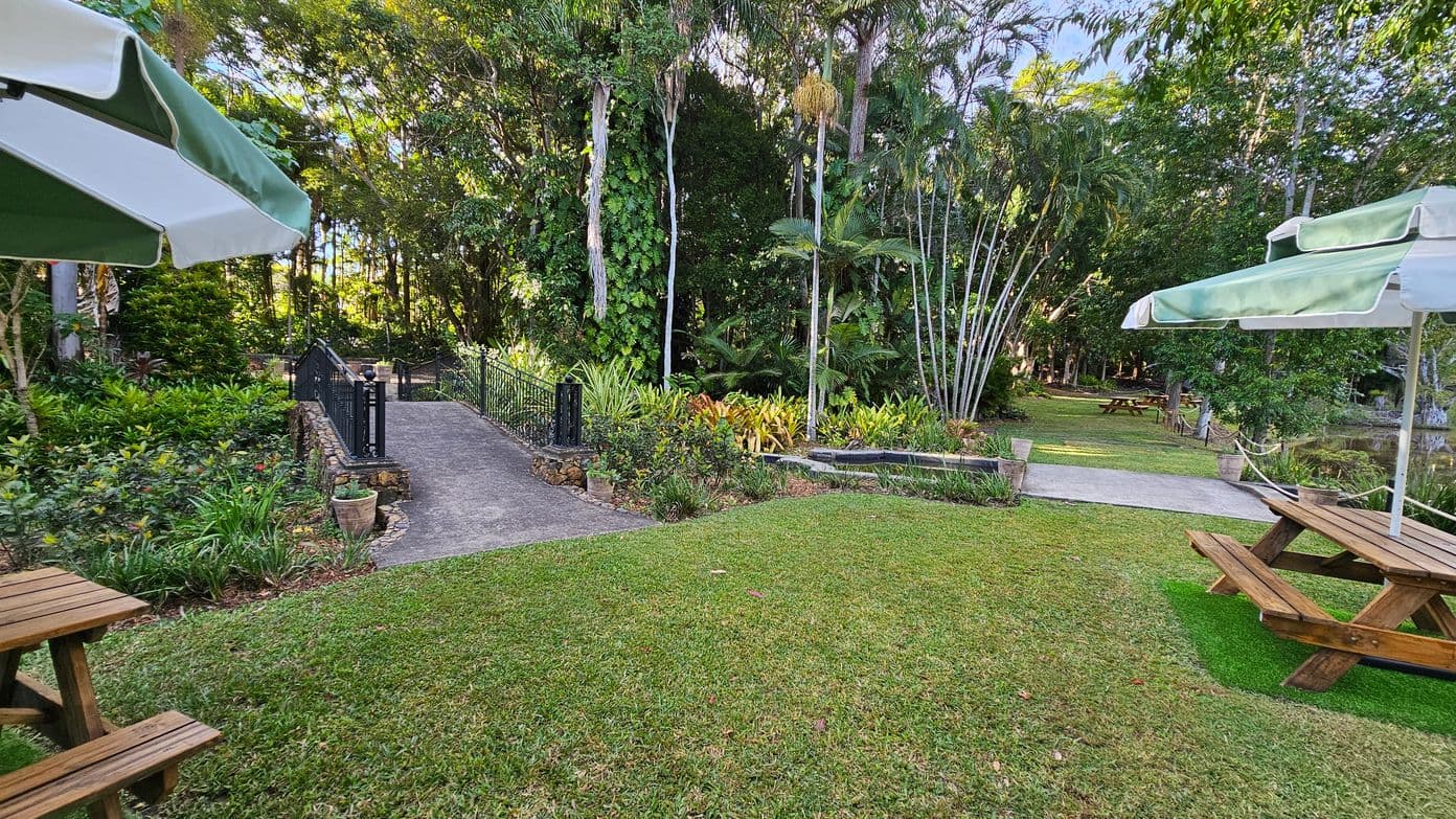 A lush garden setting with a pathway in the centre leading to a small bridge with black railings. On the left, there are dense green shrubs and trees, while on the right, tall palm trees and more greenery are visible. Two wooden picnic tables with green and white umbrellas are on either side of the image, set on a well-maintained grassy area. In the background, more trees and a glimpse of a pond can be seen.