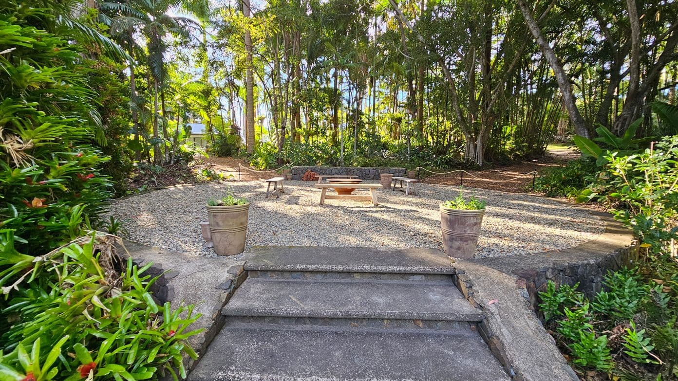 Stone steps lead up to a circular gravel area with a central fire pit surrounded by wooden benches. Two large potted plants flank the steps. Dense greenery and tall trees form a lush backdrop, with sunlight filtering through the leaves. A narrow path winds through the trees on the right. The area is bordered by low stone walls and more plants, creating a secluded, natural setting.