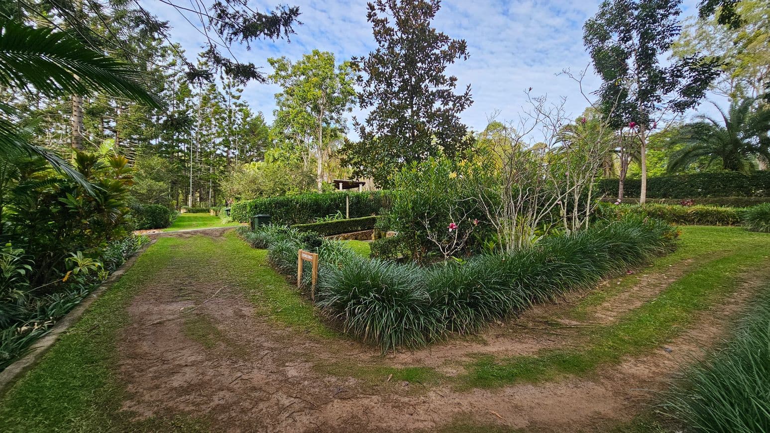 A lush garden scene with a dirt path running through the centre, bordered by green grass. On the left, dense foliage and tall trees create a natural boundary. To the right, a manicured garden area features neatly trimmed bushes and flowering plants. In the background, more trees and greenery extend into the distance, with a small structure partially visible. A wooden sign is placed near the garden area.
