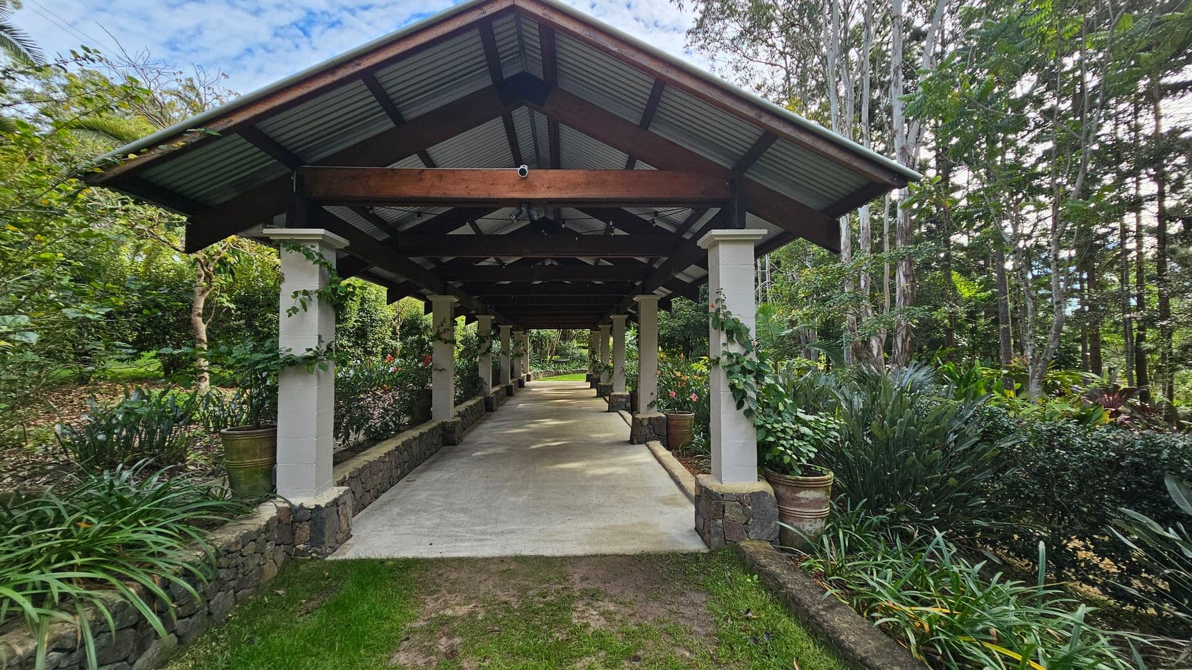 A covered walkway with a metal roof supported by stone pillars extends into the distance. The path is flanked by lush greenery and potted plants on both sides. The ground is a mix of grass and concrete, and the surrounding area is filled with tall trees and dense foliage, creating a serene, natural environment.