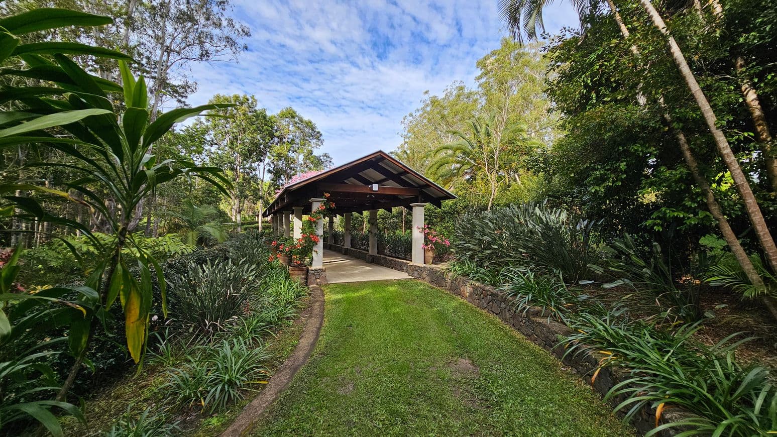 A pathway leads to a covered structure with a pitched roof supported by columns. The path is bordered by lush greenery and flowering plants in pots. On the left, tall trees and dense foliage create a natural backdrop. On the right, more trees and shrubs add to the verdant setting. The ground is covered with grass, and the structure is surrounded by a low stone wall.