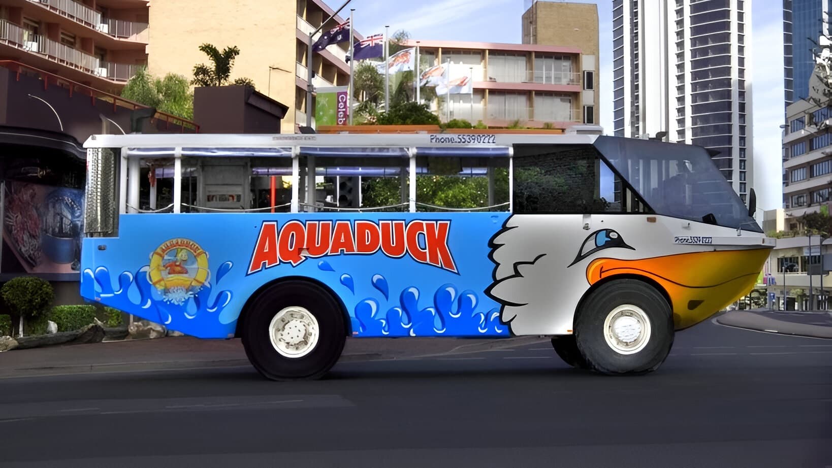 A colourful amphibious vehicle with the name "Aquaduck" in bold red letters on the side. The vehicle has a blue and white design with a cartoon duck face near the front. It is parked on a city street with tall buildings in the background, including a high-rise with a modern design. There are flags and greenery visible, and the road is smooth and dark.