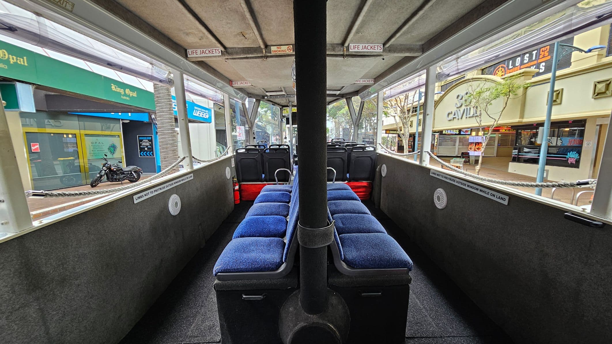 Inside a vehicle with blue cushioned seats arranged in two rows along the centre aisle. A black pole runs vertically in the middle. The ceiling is grey with signs for life jackets and no smoking. Large windows on both sides show a street view with shops, including "King Opal" on the left and "Lost" on the right. A motorcycle is parked outside on the left. The floor is carpeted in dark grey.