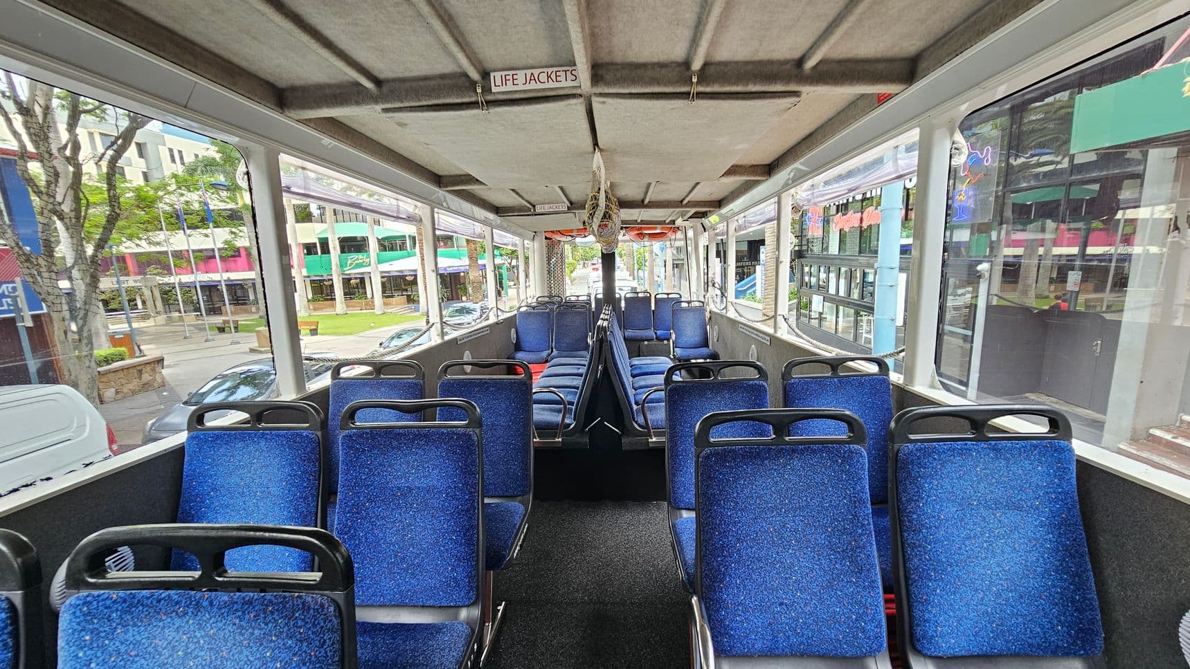 Interior of a bus with blue upholstered seats arranged in rows on both sides of a central aisle. The ceiling has a sign reading "LIFE JACKETS" and life jackets are visible. Large windows on both sides provide a view of the street outside, showing buildings, trees, and parked cars. The floor is carpeted in dark material. The bus appears to be stationary, with no passengers present.