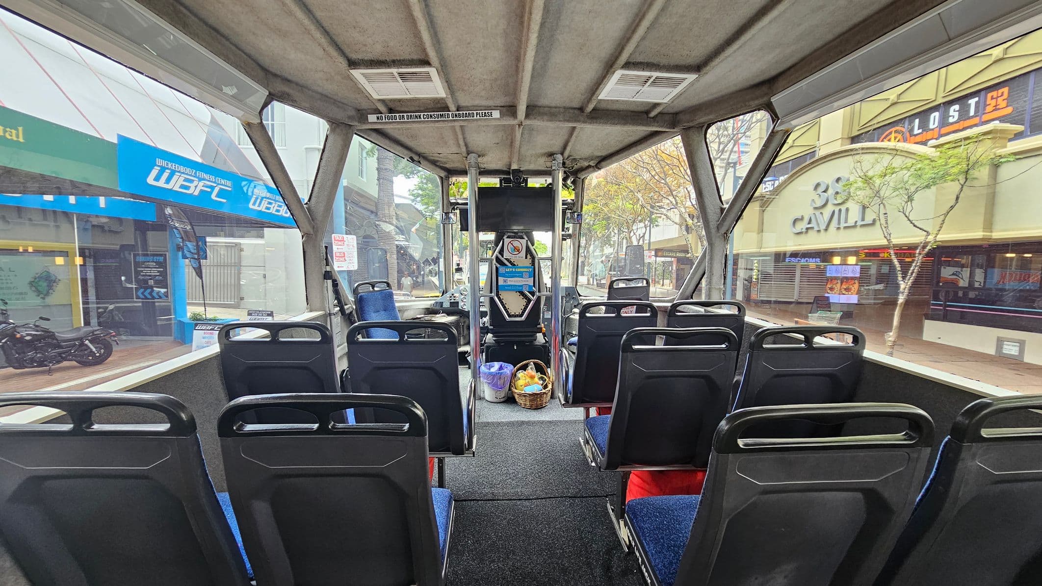 Inside a bus with blue upholstered seats arranged in rows. The floor is carpeted in dark grey. A basket with items is placed near the front. Large windows on both sides reveal a street view with shops, including a blue sign for a fitness centre on the left and a building with "38 Cavill" on the right. The ceiling is grey with ventilation panels and a sign reading "No food or drink consumed onboard please."