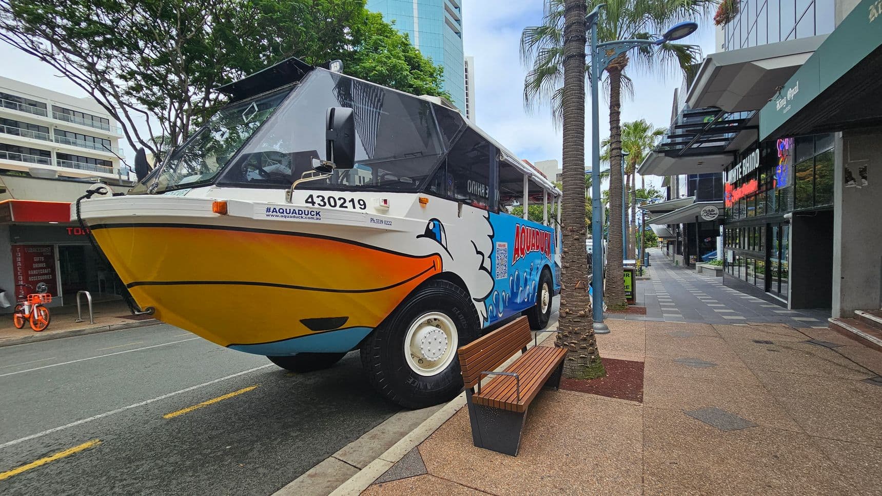 A colourful amphibious vehicle with a duck design is parked on a city street. On the left, there are trees and a building with a white facade. The vehicle has large wheels and a yellow front. On the right, a pavement lined with palm trees and modern buildings, including a place named "WHITE RHINO". A wooden bench is positioned near the vehicle. The street is marked with yellow lines, and a bright orange bicycle is visible in the background.