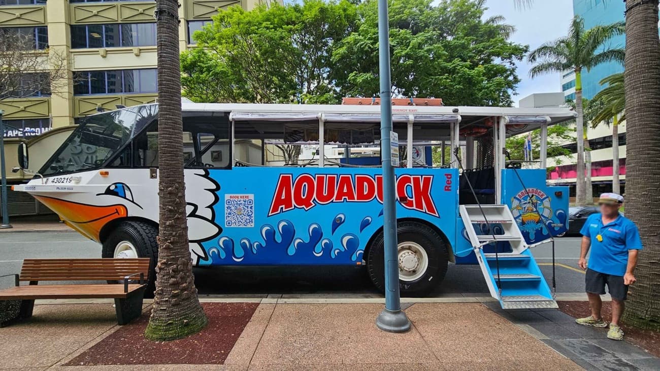 A colourful amphibious vehicle with "Aquaduck" written in bold red letters on a blue background is parked on a street. The vehicle has a cartoon duck design on the front. A set of steps leads up to the entrance. A person in a blue uniform and captain's hat stands to the right. The background features a building with green accents and several palm trees. A wooden bench is on the left, and a streetlamp is in the centre.