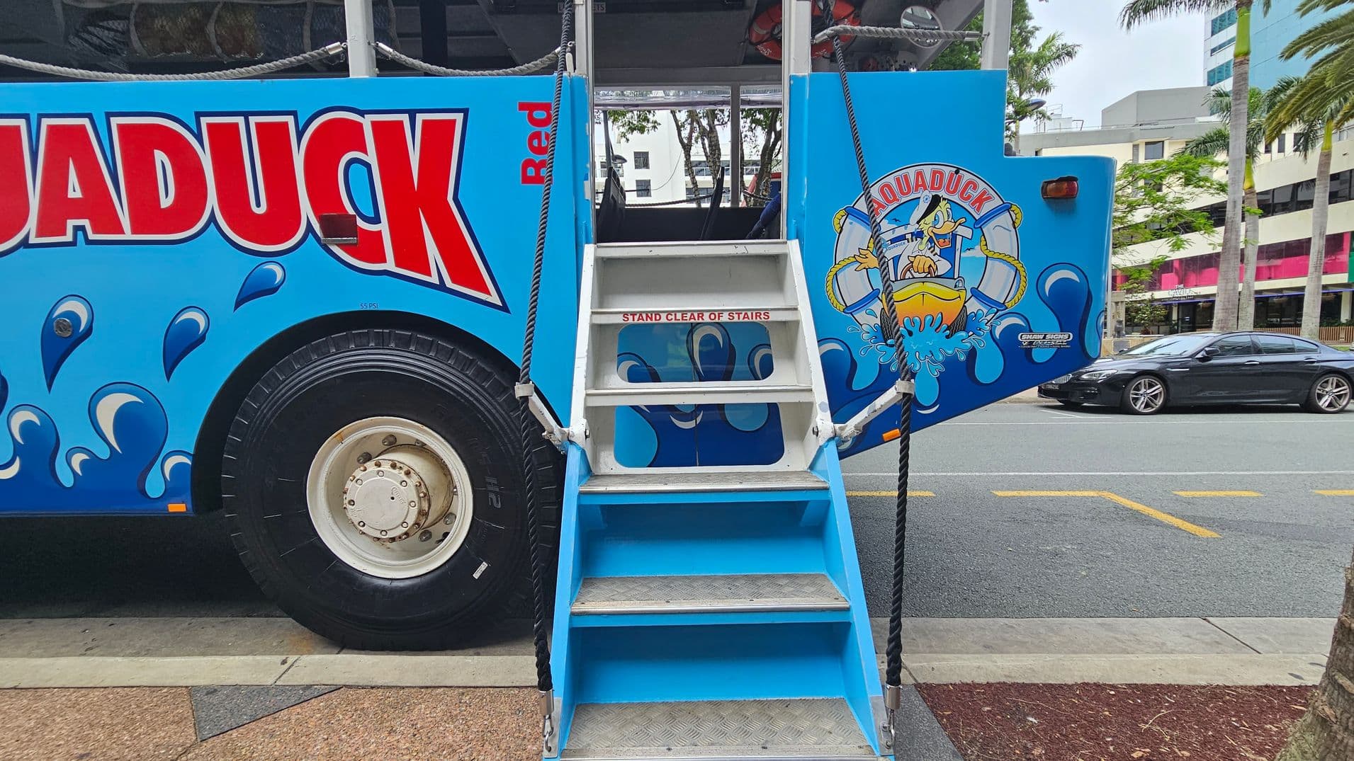 On the left, a large blue vehicle with the word "QUADDUCK" in bold red letters and water splash graphics. In the centre, a set of blue and white stairs with a sign reading "STAND CLEAR OF STAIRS" leads into the vehicle. On the right, a cartoon duck in a life preserver is depicted on the vehicle's side. In the background, a street with a black car, palm trees, and buildings. The ground is a mix of concrete and textured surfaces.
