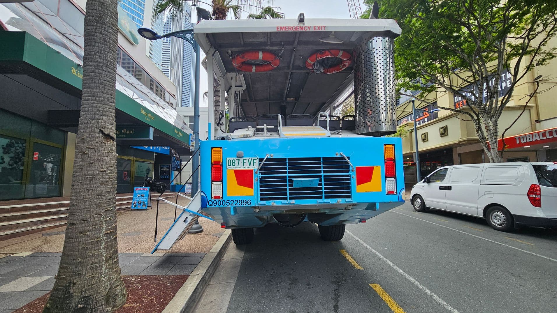 A large blue amphibious vehicle is parked on a city street. It has an open top with life rings visible inside and an "Emergency Exit" sign above. The vehicle's rear features a licence plate and various lights. On the left, there's a palm tree and a building with green awnings. On the right, a white van is parked, and a building with signs is visible. The street is lined with trees and has a pavement area with steps leading to shops.