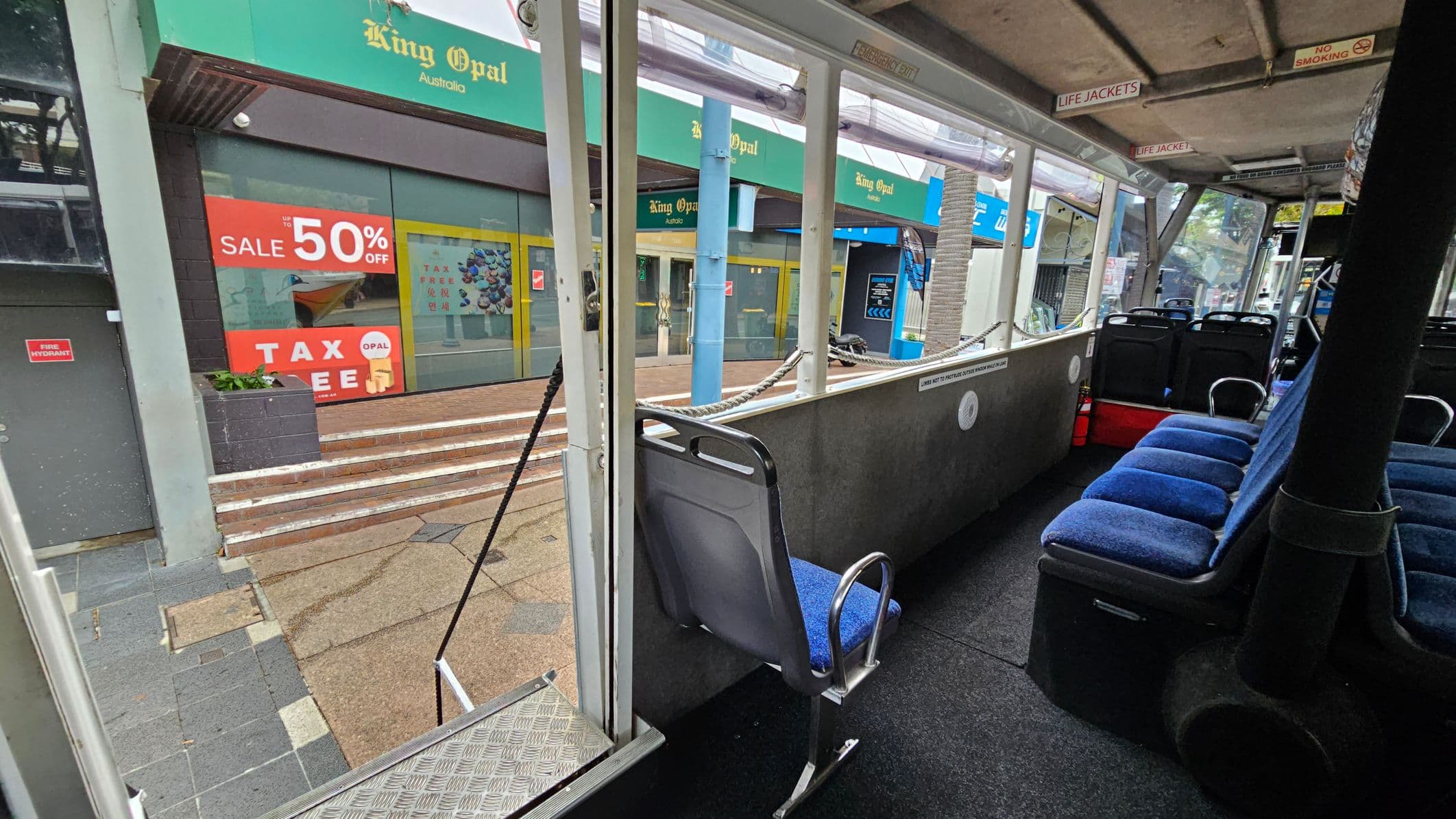 Inside a bus with blue cushioned seats on the right. A single seat is near the open door on the left. The floor is dark and textured. The ceiling has signs for life jackets and no smoking. Outside, a shop with green signage reads "King Opal Australia" and displays a large red sale sign offering 50% off. Steps lead up to the shop entrance, and a fire hydrant sign is visible on a grey wall. The pavement outside is a mix of grey and beige tiles.