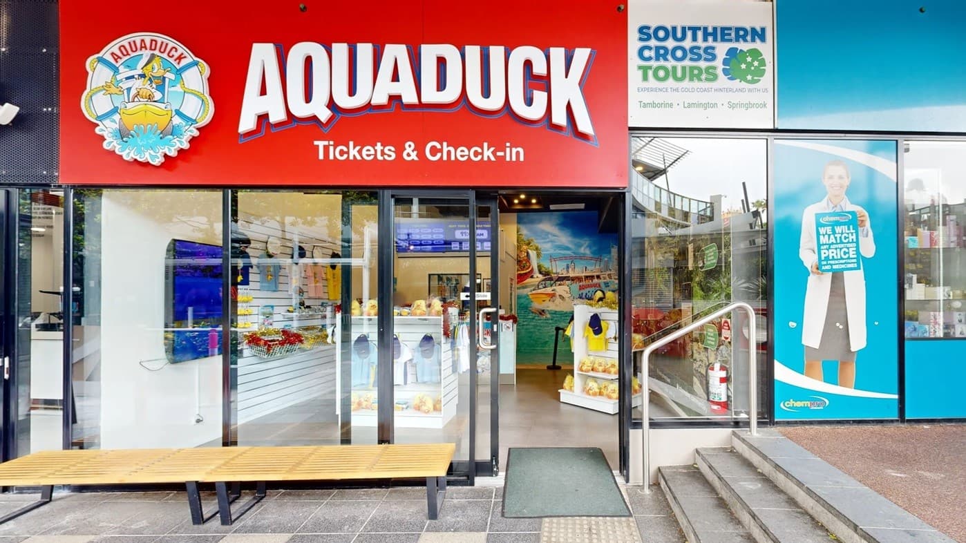On the left, a red sign with "AQUADUCK Tickets & Check-in" and a logo above a glass entrance. Inside, there are displays with merchandise and a mural of a boat on water. On the right, a blue sign for "SOUTHERN CROSS TOURS" and a large poster of a person in a white coat holding a sign. A set of steps with a metal railing leads up to the entrance. A wooden bench is in front of the glass doors, and the floor is tiled.