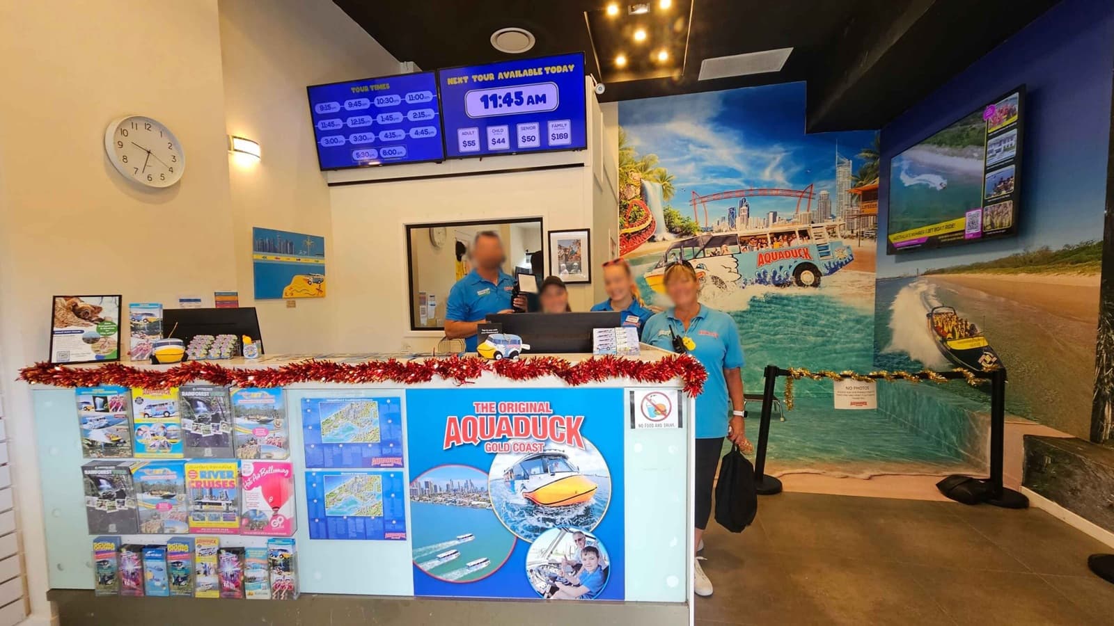A reception area with four people in blue shirts standing behind a counter decorated with red tinsel. The counter displays brochures and a sign for "The Original Aquaduck Gold Coast." On the left wall, a clock shows the time as 11:45. Above the counter, digital screens display tour times and prices. The right wall features a large mural of a beach scene with an Aquaduck vehicle. The floor is tiled, and the ceiling has recessed lighting.