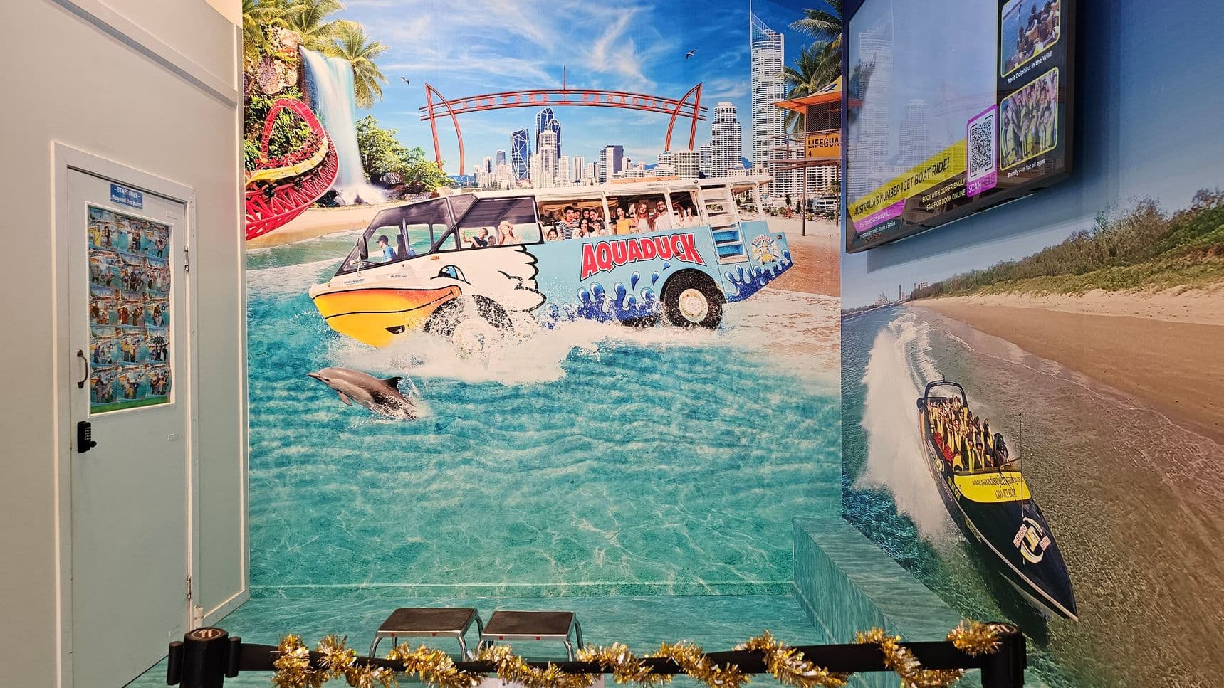A vibrant mural covers the walls, depicting a lively aquatic scene. On the left, a door with a collage of images. In the centre, a colourful amphibious vehicle labelled "Aquaduck" splashes through water, with a dolphin nearby. The background features a city skyline, a waterfall, and a roller coaster. On the right, a speedboat with passengers rides along a beach. A screen displaying information is mounted above. The floor has a water-themed design, and a garland decorates the foreground.