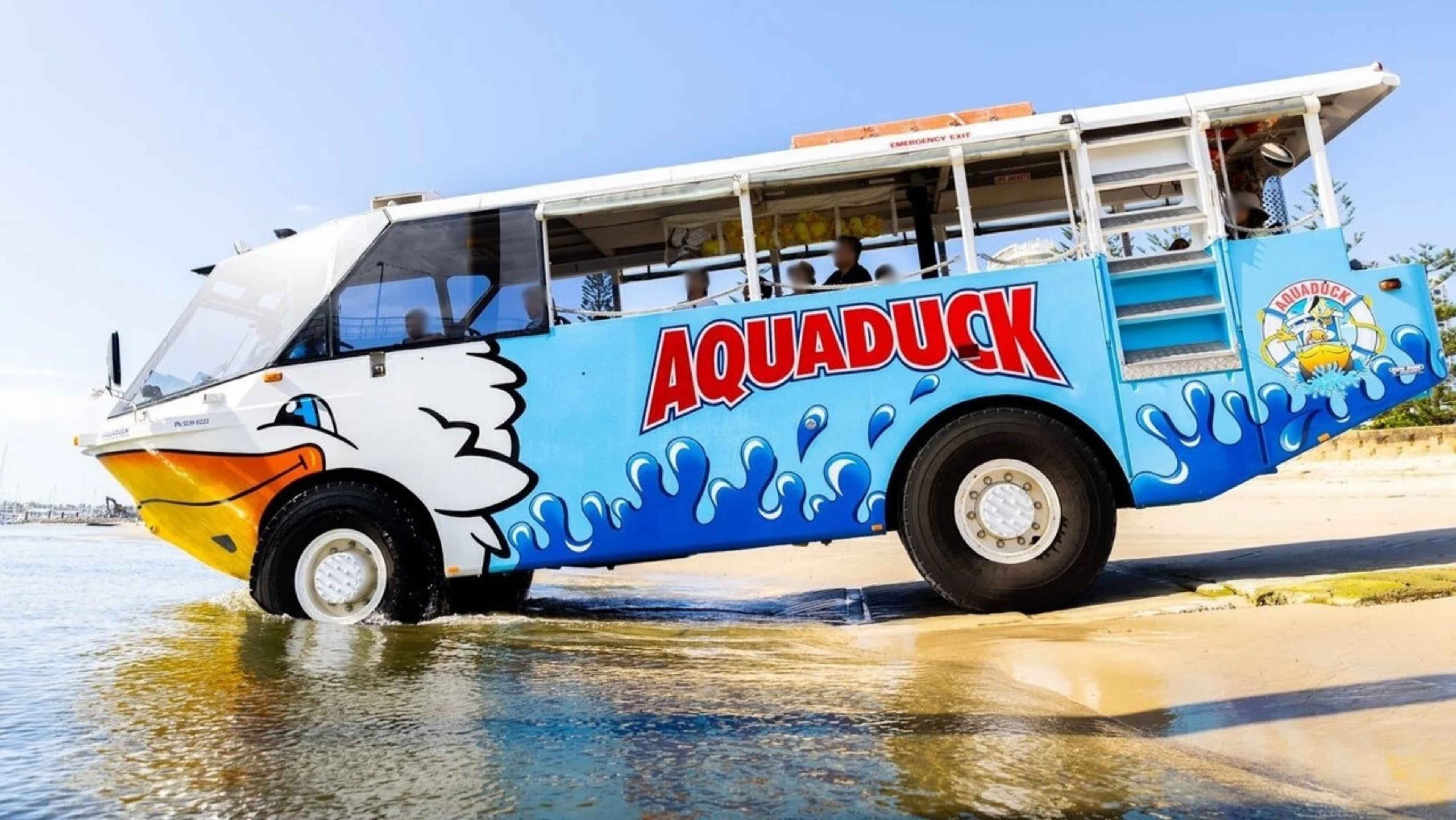 An amphibious vehicle with the name "Aquaduck" painted on the side is partially in the water. The vehicle is designed with a duck motif, featuring a white and orange front resembling a duck's beak. The body is blue with wave patterns and a logo on the side. Several passengers are visible inside. The vehicle is on a sandy beach, with water in the foreground and a clear sky in the background.